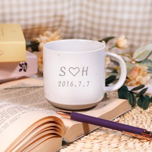 May include: White ceramic mug with a brown base, featuring the initials "S ♡ H" and the date "2016.7.7". The mug is placed on a book, with a purple pen and other items in the background.