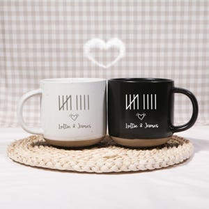 Puede incluir: Dos tazas de cerámica, una blanca y otra negra, con un diseño minimalista. Cada taza presenta líneas verticales, un corazón y los nombres "Lottie & James". Las tazas están sobre un posavasos tejido.