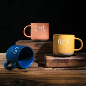 May include: Three ceramic mugs in orange, yellow, and blue, each with a handle. The orange and yellow mugs have the word "OMA" printed on them, while the blue mug has "OPA". All mugs have a heart symbol and the text "EST 2026".