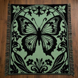 May include: A rectangular crochet blanket featuring a large, central butterfly motif in black against a light mint green background. The design is framed by an arched border with floral accents and leaves, finished with short fringe along the sides.