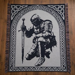 May include: A rectangular crochet blanket featuring a black and white graphic of a knight kneeling and holding a sword. The design is enclosed within an arched frame decorated with intricate Celtic knotwork patterns and finished with fringed edges.