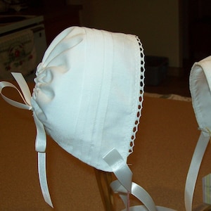 May include: Two white bonnets with white satin ribbons. The bonnets are displayed on gold stands.