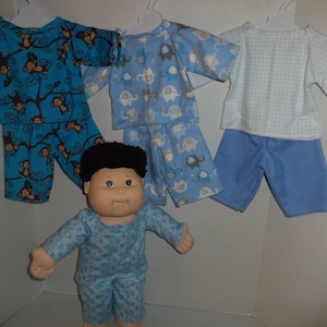 May include: Three sets of pajamas for a doll, each with a different pattern. One set features monkeys on a blue background, another has elephants on a light blue background, and the third has a white and blue gingham pattern. A doll wearing a blue and white patterned pajama set is standing in front of the pajamas.