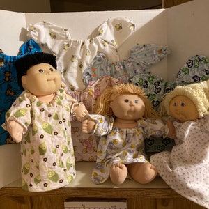 May include: Five Cabbage Patch Kids dolls wearing various patterned pajamas. The dolls are sitting on a wooden shelf with a white background.