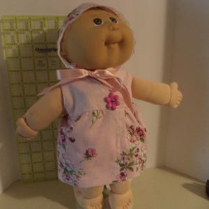 May include: A Cabbage Patch Kid doll wearing a pink floral dress with a pink satin bow and a pink flower accent. The doll has a light brown complexion and brown eyes.