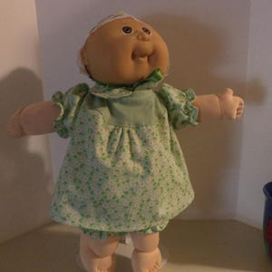 May include: A Cabbage Patch Kid doll wearing a green floral dress with a white collar and a white headband. The doll has a light brown complexion and brown eyes.