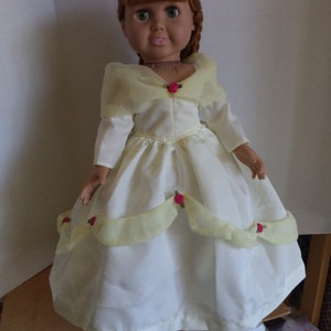 May include: A doll wearing a white and yellow dress with pink rose accents. The dress has a long skirt and a fitted bodice. The doll has red hair and blue eyes.