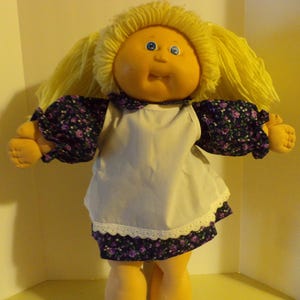 May include: A Cabbage Patch Kid doll with blonde hair, blue eyes, and a white apron over a purple and black floral dress.