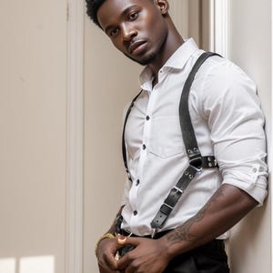 May include: A white shirt, black trousers, and black leather suspenders with metal buckles. The suspenders are attached to the trousers. The person is leaning against a wall. The image is well-lit.