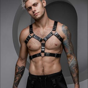 May include: Black leather body harness with silver-tone metal rings and rivets. The harness features adjustable straps and a Y-shaped design across the chest. The product is worn over the torso.