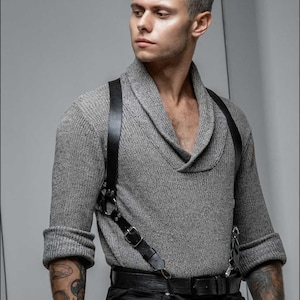 May include: A grey knitted sweater with a shawl collar and rolled-up sleeves. The outfit includes a black leather harness and belt, paired with black trousers. The model has tattoos on their arm.
