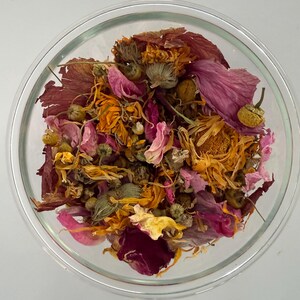 May include: A clear glass bowl filled with a colorful assortment of dried flowers and petals. The mix includes shades of orange, pink, and burgundy, with some green buds. The arrangement is set against a plain white background.