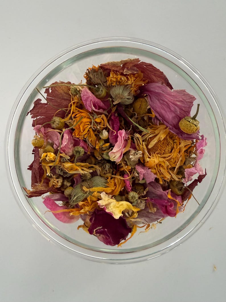 May include: A clear glass bowl filled with a colorful assortment of dried flowers and petals. The mix includes shades of orange, pink, and burgundy, with some green buds. The arrangement is set against a plain white background.