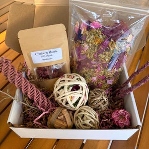 May include: A gift box filled with natural pet toys and treats. The box contains a bag of dried flowers, a bag of cranberry hearts, woven balls, and dried floral accents. The box is white and sits on a wooden surface.
