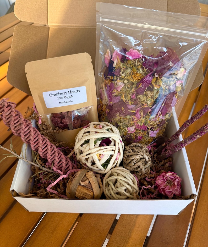 May include: A gift box filled with natural pet toys and treats. The box contains a bag of dried flowers, a bag of cranberry hearts, woven balls, and dried floral accents. The box is white and sits on a wooden surface.