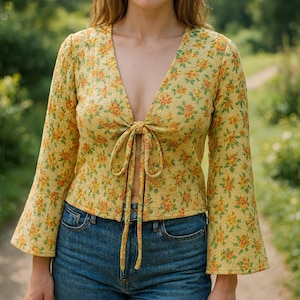 May include: A yellow floral top with a V-neckline and tie closure. The long-sleeved top features a pattern of orange and green flowers. The model is wearing blue jeans. The background is a blurred outdoor scene.