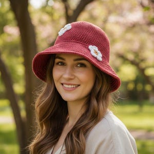 May include: A claret-coloured crocheted sun hat with a wide brim and white flower accents. The hat features two white flower appliqués on the side. This handmade hat is designed for sun protection and adds a touch of charm.