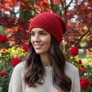 Könnte beinhalten: Eine Frau trägt eine rote Strickmütze und einen cremefarbenen Pullover. Die Mütze hat eine einfarbige, tiefrote Farbe und scheint aus einem groben Strick gefertigt zu sein. Der Hintergrund zeigt rotes und grünes Laub und rote Rosen.