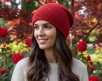 May include: A woman wearing a red knitted beanie and a cream-colored sweater. The beanie is a solid, deep red color and appears to be made of a chunky knit. The background features red and green foliage and red roses.