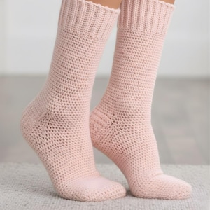 Könnte beinhalten: Ein Paar hellrosa Häkelsocken. Die Socken sind kniehoch mit geripptem Bündchen und einem strukturierten, offen gewebten Design. Die Socken werden auf einer hellen Oberfläche präsentiert.