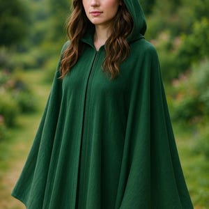 Pode incluir: Uma capa com capuz verde escuro com um design longo e fluido. A capa tem um capuz e um fecho frontal. O tecido parece ser um material macio e texturizado. A pessoa está usando a capa ao ar livre.