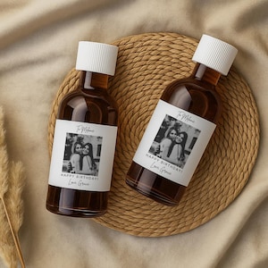 May include: Two glass bottles filled with a dark liquid, each with a white label featuring a black and white photo and text. The labels read "To Melanie, HAPPY BIRTHDAY! Love, Grace." The bottles have white caps and are on a woven mat.