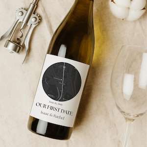 May include: A wine bottle with a custom label that reads "OUR FIRST DATE" and the date June 16, 1999, with the names Isaac & Rachel. The label features a black and white map design. A wine glass and corkscrew are also visible.