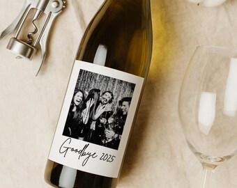 May include: A wine bottle with a label featuring a black and white photo of people. The label reads "Goodbye 2025" in a handwritten font. A silver corkscrew and a wine glass are next to the bottle.