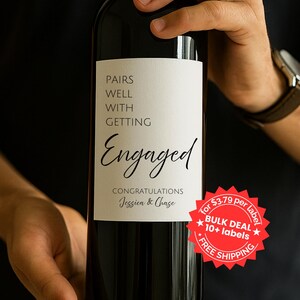 May include: A dark red wine bottle with a white label that reads "PAIRS WELL WITH GETTING Engaged". The label also includes "CONGRATULATIONS Jessica & Chase" in script. The bottle is held by a person.
