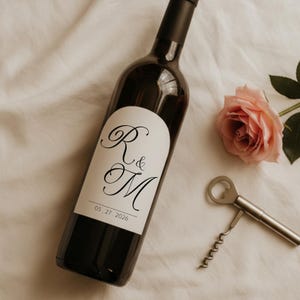 May include: A dark red wine bottle with a white label featuring the initials "R & M" in elegant script, and the date "05 27 2026". A silver corkscrew and a pink rose are next to the bottle. The bottle is on a white cloth.