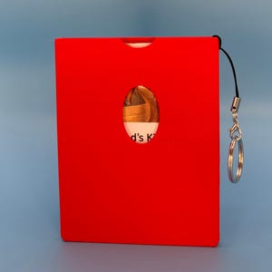May include: A bright red rectangular card holder with a small oval cutout revealing a glimpse of the contents. The holder has a black lanyard with a silver-colored charm and a key ring attached. The text "d's K" is visible through the cutout.