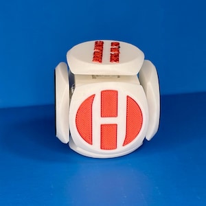 Puede incluir: Objeto cúbico blanco con una "H" roja en una cara y dos barras verticales rojas en la parte superior. El objeto tiene un diseño geométrico moderno y parece impreso en 3D. El fondo es azul liso.