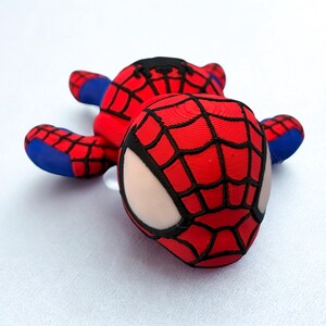 May include: A red and black Spider-Man plush toy with a web pattern on the head and body. The toy has blue accents on the arms and feet, and white eye details. The plush is lying on a white surface.