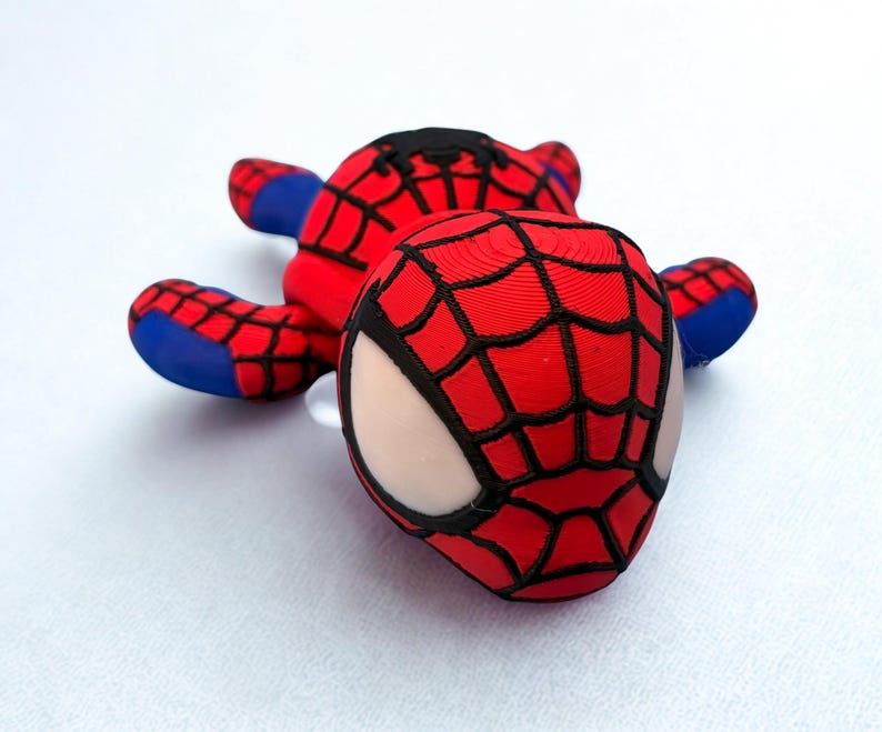 May include: A red and black Spider-Man plush toy with a web pattern on the head and body. The toy has blue accents on the arms and feet, and white eye details. The plush is lying on a white surface.