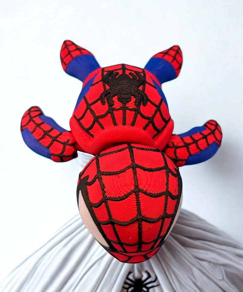 May include: A red and blue Spider-Man-themed plush toy. The toy features a red body with black web detailing and a black spider emblem. The ears and accents are blue. The toy is resting on a white surface.