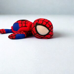 May include: A red, blue, and black Spider-Man figurine. The figure is lying down, with the head and body visible. The head is red with a black web pattern and white eye shapes. The body is red and blue.