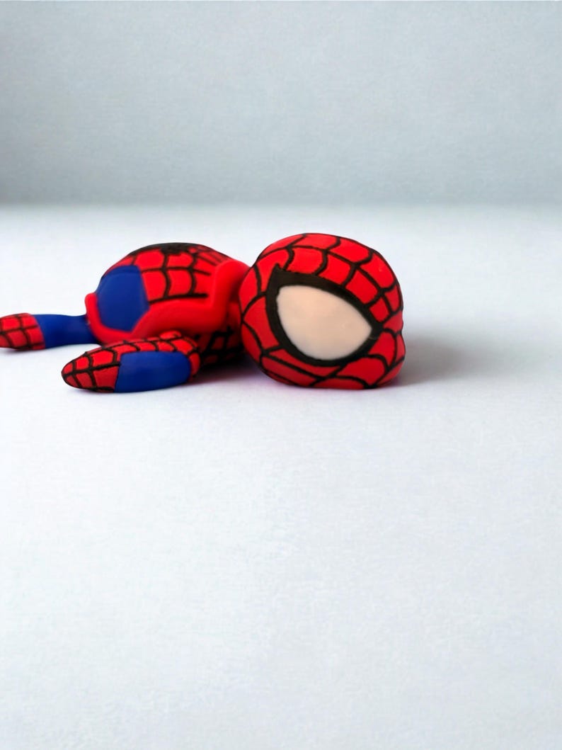 May include: A red, blue, and black Spider-Man figurine. The figure is lying down, with the head and body visible. The head is red with a black web pattern and white eye shapes. The body is red and blue.