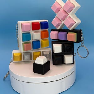 May include: A collection of colorful, cube-shaped keychains in various arrangements. The keychains feature a variety of colors, including pink, blue, white, and yellow. Some are arranged in a grid, while others form an X shape. Each keychain has a silver keyring.