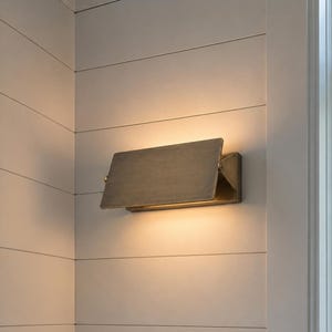 Könnte beinhalten: Eine rechteckige Wandleuchte mit metallischem Bronzefinish und einer klappbaren Frontplatte, die das Licht nach oben und unten lenkt und einen warmen Schein auf die weiße, horizontal getäfelte Wand im Hintergrund wirft.