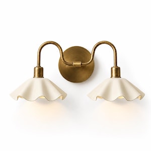 May include: A double wall sconce featuring a circular brass backplate and two curved brass arms. Each arm supports a cream-coloured, ruffled, bell-shaped shade. The fixture emits a soft, warm glow from beneath the two downward-facing shades.