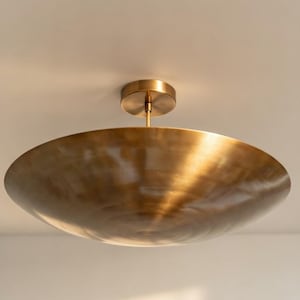 Könnte beinhalten: Eine messingfarbene Deckenleuchte mit einem flachen, schalenförmigen Schirm. Die Leuchte ist bündig an der Decke montiert und hat einen zentralen Stiel und eine kreisförmige Basis. Das Metall hat eine warme, reflektierende Oberfläche.