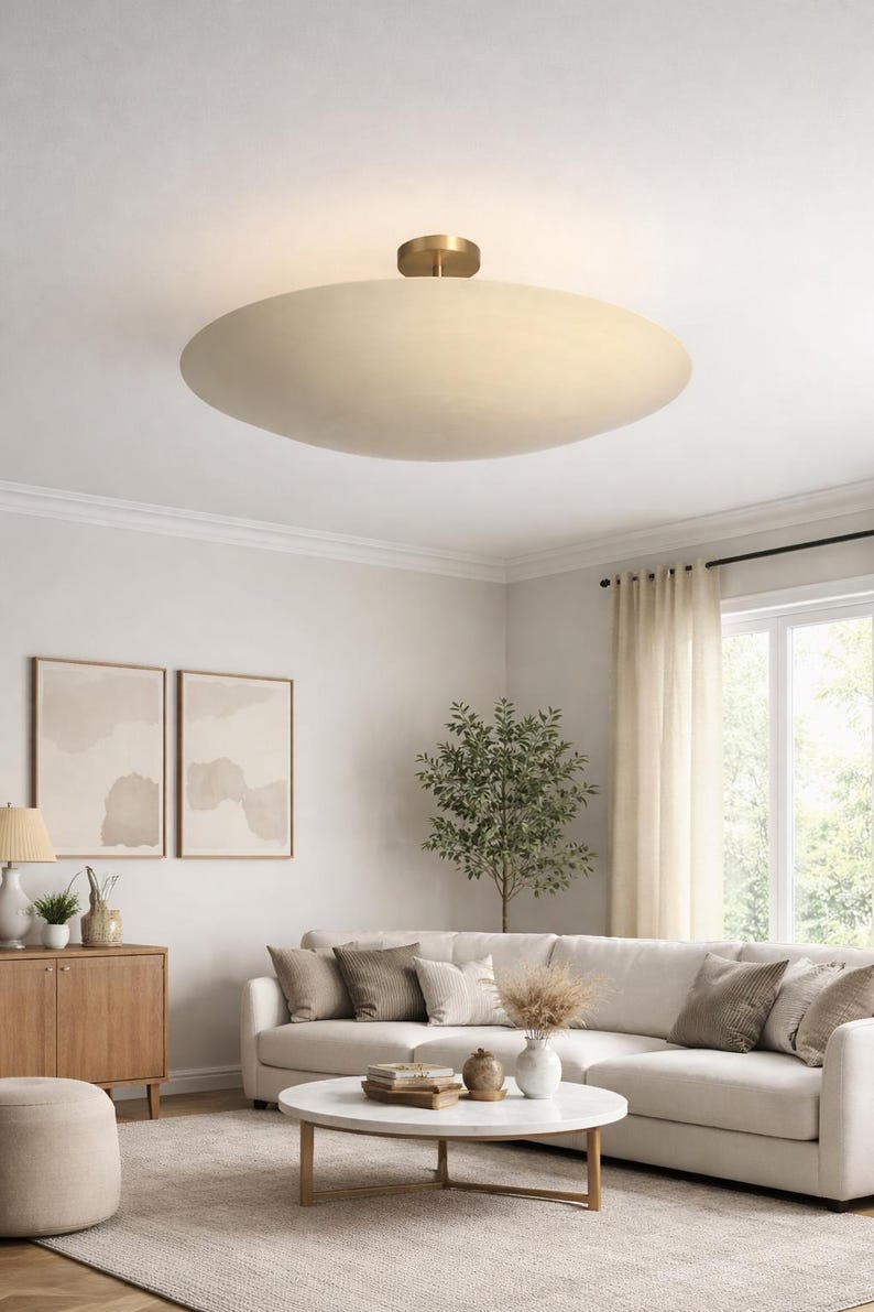 May include: A contemporary, cream-coloured, oval ceiling light with a brass central mount. The light illuminates a neutral-toned living room, featuring a sectional sofa, coffee table, and artwork. The room has a modern aesthetic.