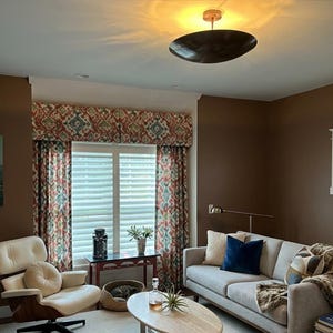 K&ouml;nnte beinhalten: Ein Wohnzimmer mit modernem Design. Zu den Merkmalen geh&ouml;ren ein hellgraues Sofa mit Wurfkissen und einer Kunstfelldecke, ein cremefarbener Stuhl und gemusterte Vorh&auml;nge. Eine runde, dunkel gef&auml;rbte Leuchte h&auml;ngt von der Decke.