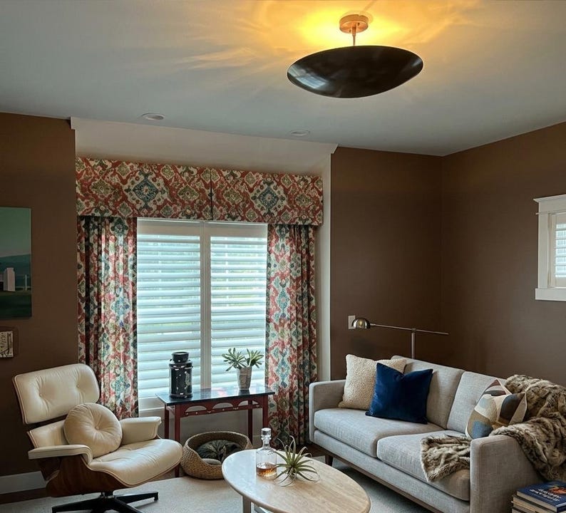 K&ouml;nnte beinhalten: Ein Wohnzimmer mit modernem Design. Zu den Merkmalen geh&ouml;ren ein hellgraues Sofa mit Wurfkissen und einer Kunstfelldecke, ein cremefarbener Stuhl und gemusterte Vorh&auml;nge. Eine runde, dunkel gef&auml;rbte Leuchte h&auml;ngt von der Decke.