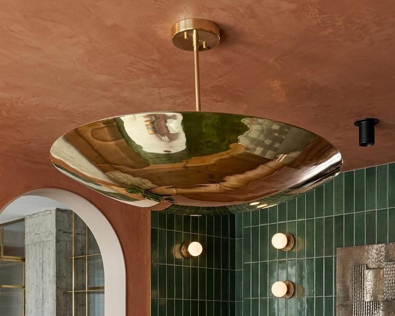 K&ouml;nnte beinhalten: Eine gro&szlig;e, goldfarbene, schalenf&ouml;rmige Deckenleuchte, die an einer bronzefarbenen Stange und Deckenhalterung aufgeh&auml;ngt ist. Das Licht reflektiert die Umgebung. Der Hintergrund zeigt eine gr&uuml;ne geflieste Wand und eine rotbraune Decke.