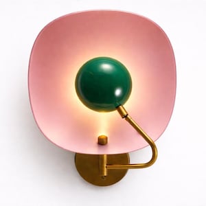 Puede incluir: Aplique de pared moderno con una pantalla rosa redondeada y una esfera de luz verde. El accesorio tiene un brazo y una base dorados, creando un contraste elegante. La luz emite un brillo cálido, realzando el diseño. Medidas en cm.