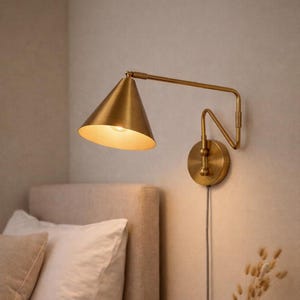 Può includere: Applique da parete color ottone con paralume conico e braccio regolabile. L'applique è montata a parete e ha un cavo di alimentazione visibile. La luce è accesa, illuminando l'interno del paralume. L'applique è vicino a un letto con cuscini.
