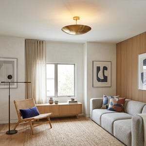 K&ouml;nnte beinhalten: Modernes Wohnzimmer mit hellgrauem Sofa, Korbstuhl und Holzschrank. Eine gro&szlig;e Deckenleuchte aus Messing und gerahmte abstrakte Kunstwerke erg&auml;nzen das moderne Design. Der Raum zeichnet sich durch eine neutrale Farbpalette mit nat&uuml;rlichen Texturen aus.