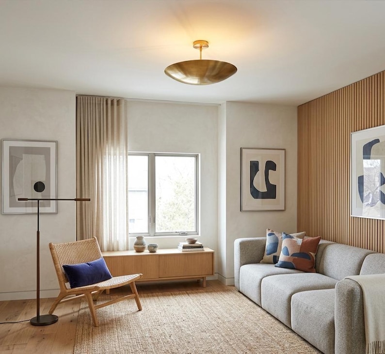 K&ouml;nnte beinhalten: Modernes Wohnzimmer mit hellgrauem Sofa, Korbstuhl und Holzschrank. Eine gro&szlig;e Deckenleuchte aus Messing und gerahmte abstrakte Kunstwerke erg&auml;nzen das moderne Design. Der Raum zeichnet sich durch eine neutrale Farbpalette mit nat&uuml;rlichen Texturen aus.