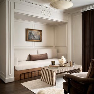 K&ouml;nnte beinhalten: Ein gem&uuml;tliches Wohnzimmer mit einem eingebauten wei&szlig;en Sofa, einem gestreiften Couchtisch und einem braunen Sessel. Der Raum verf&uuml;gt &uuml;ber ein kleines Gem&auml;lde, eine Deckenleuchte und dunkelbraune Vorh&auml;nge. Ein flauschiger wei&szlig;er Teppich liegt auf dem Boden.